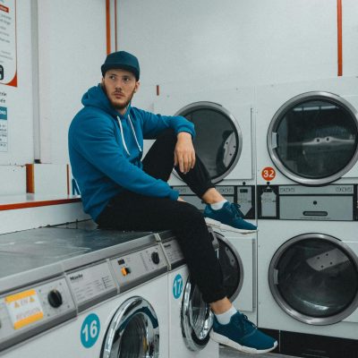 A man casually sitting in a laundromat, wearing a hoodie and cap, with washing machines in view.