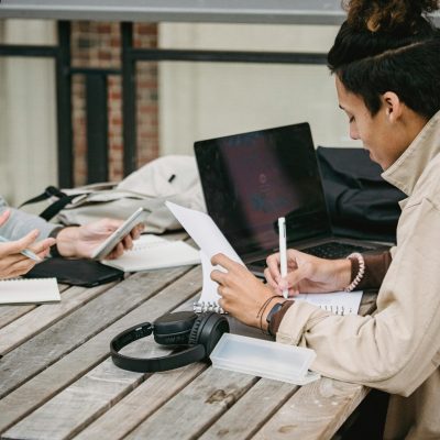 Side view of young multiracial male students in casual clothes sitting at wooden table with gadgets and taking notes while working on project