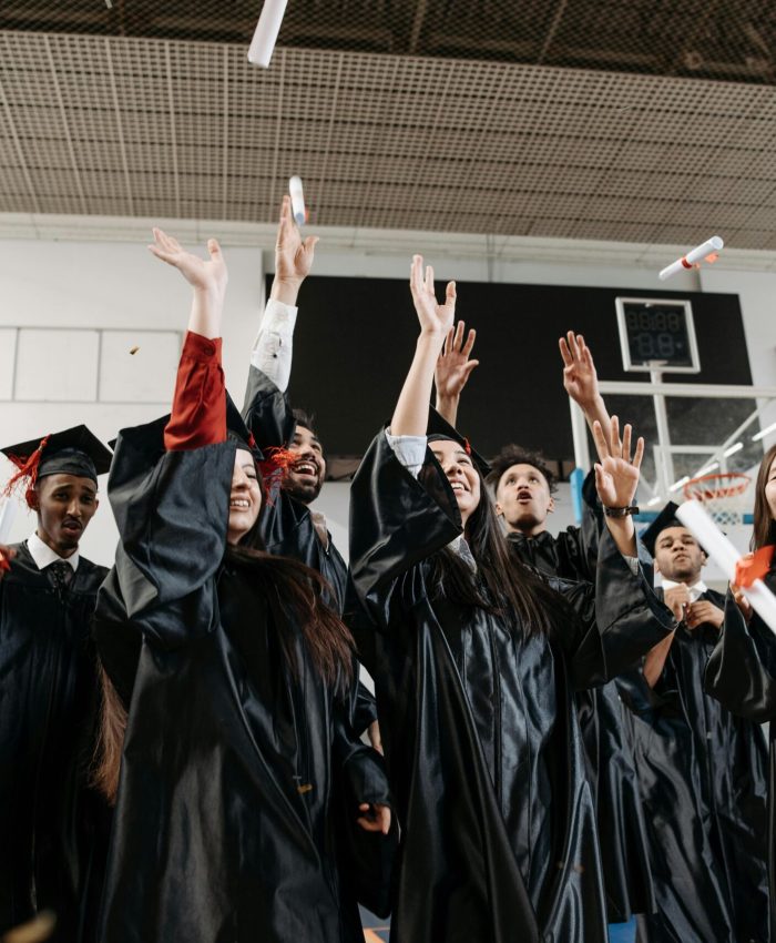 Graduation celebration with diverse students in black gowns throwing caps indoors.
